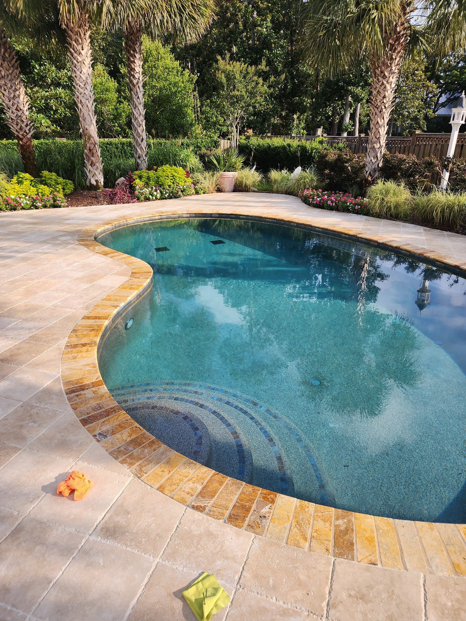 Swimming pool with tan brick border and light blue water, surrounded by landscaping, trees, and pavers.