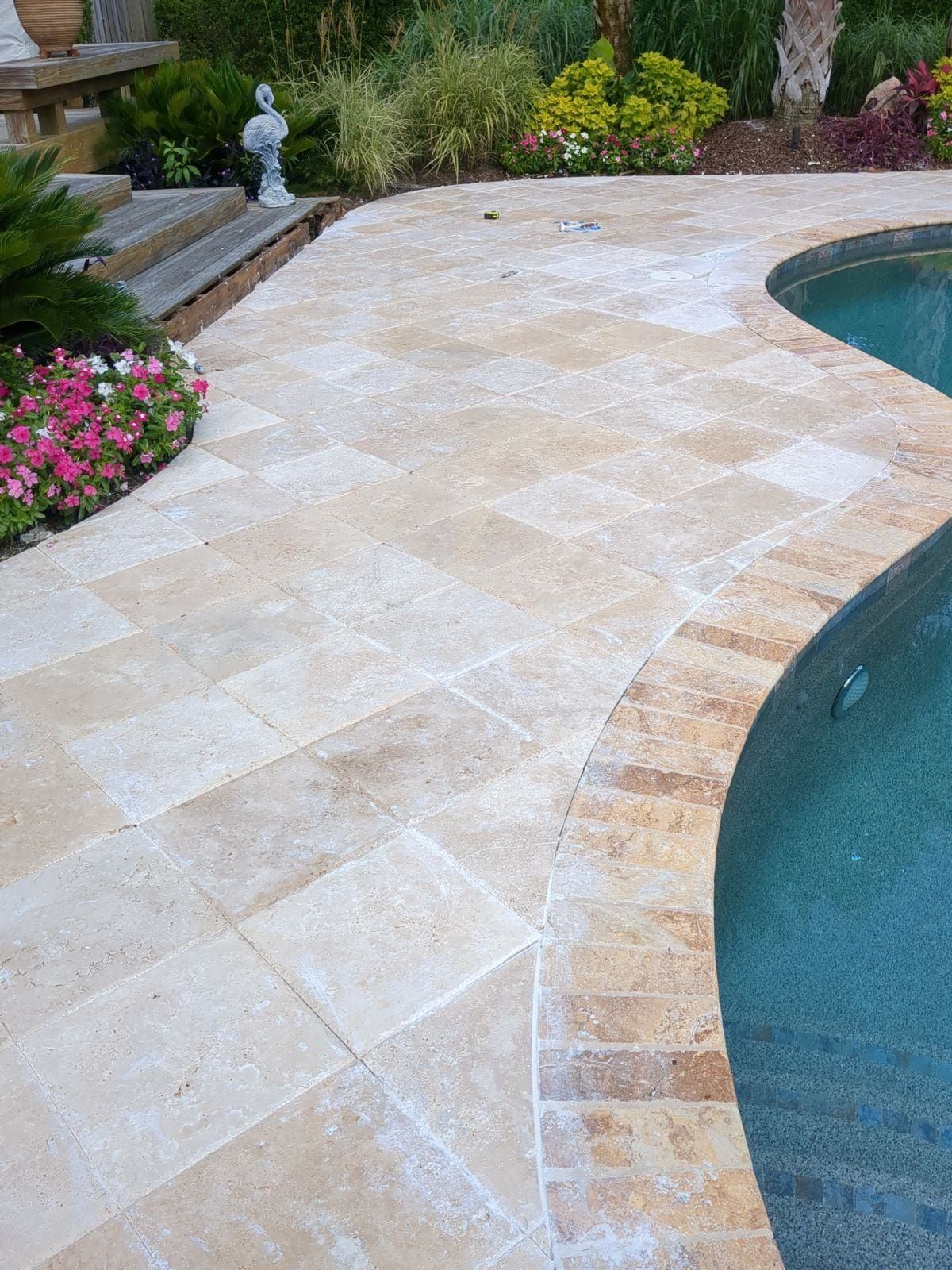 Stone patio curves around a turquoise pool. Flowers and greenery border the patio.
