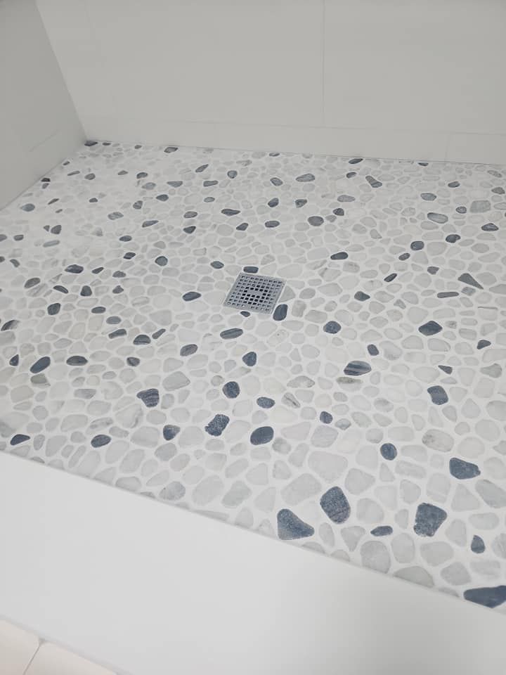 Shower floor with small, pebble-like tiles in shades of white, gray, and dark blue; drain cover visible.