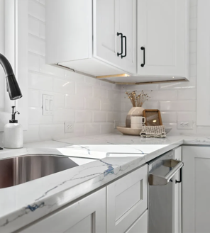Bright white kitchen corner with marble countertop, stainless sink, and white cabinets against subway tile backsplash
