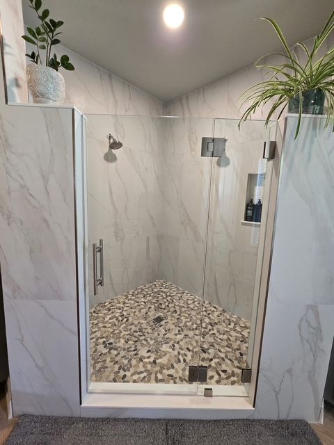 Walk-in shower with marble-look tile, pebble floor, glass door, and plants on top corners