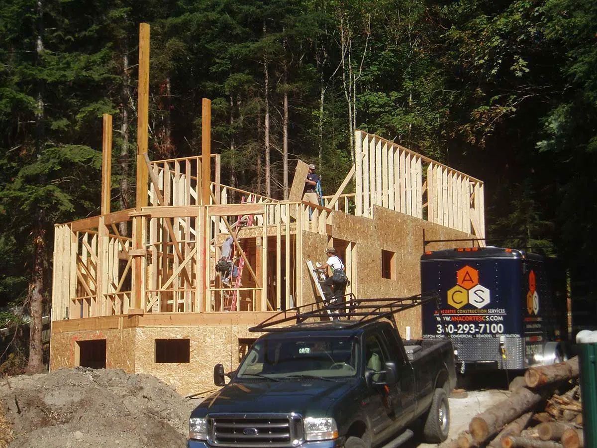 House under construction with wooden framing, workers on site, and a truck in front of a wooded area