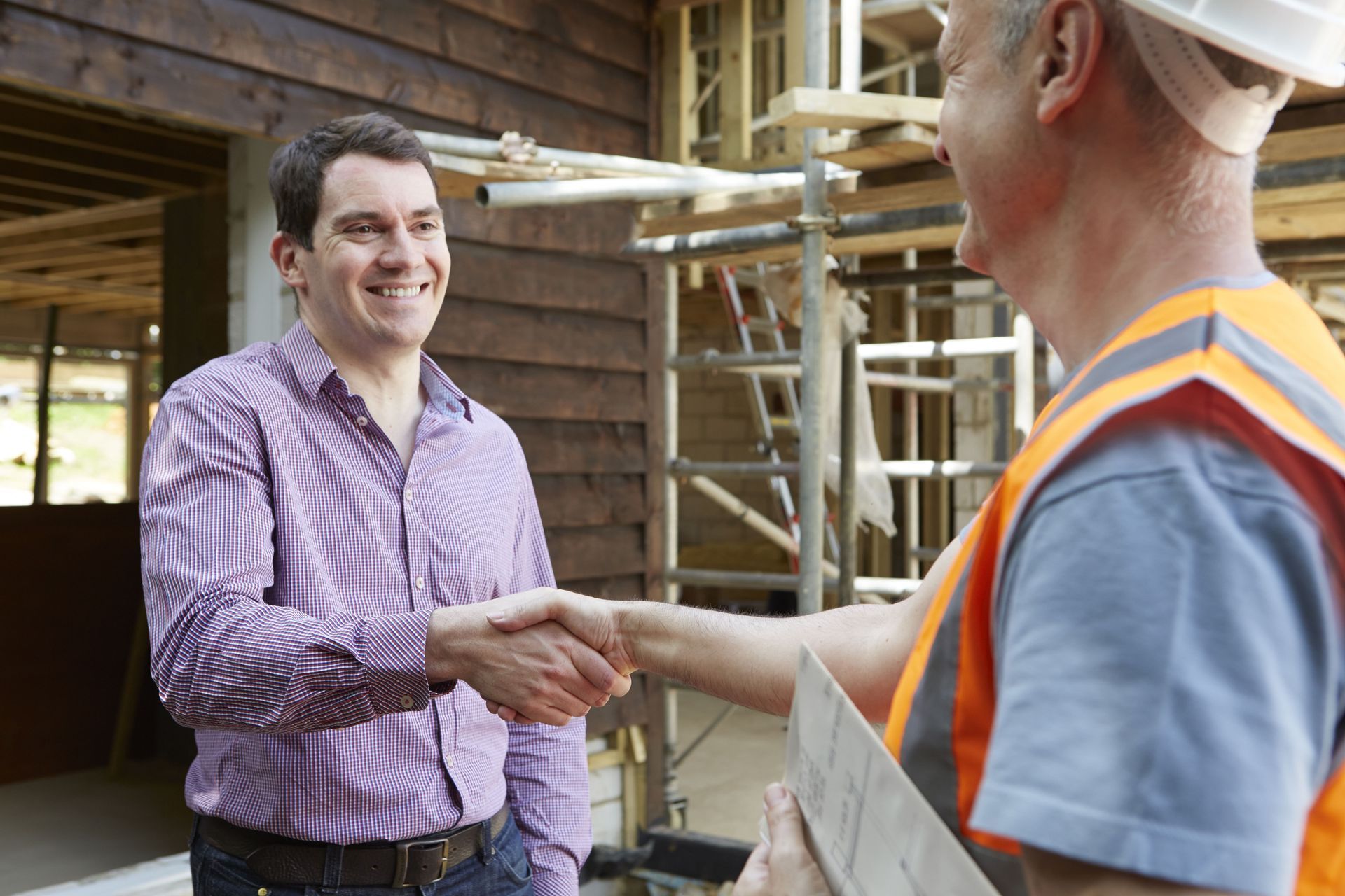 Two men shaking hands at a construction site, one in a plaid shirt and one in a safety vest.