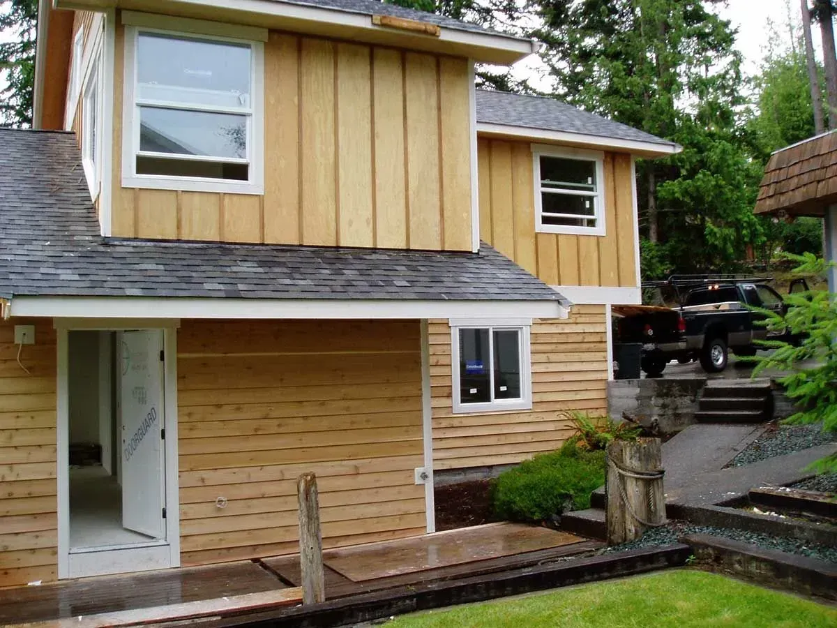 Two-story house with tan siding and a white front door, viewed from a wet driveway after rain