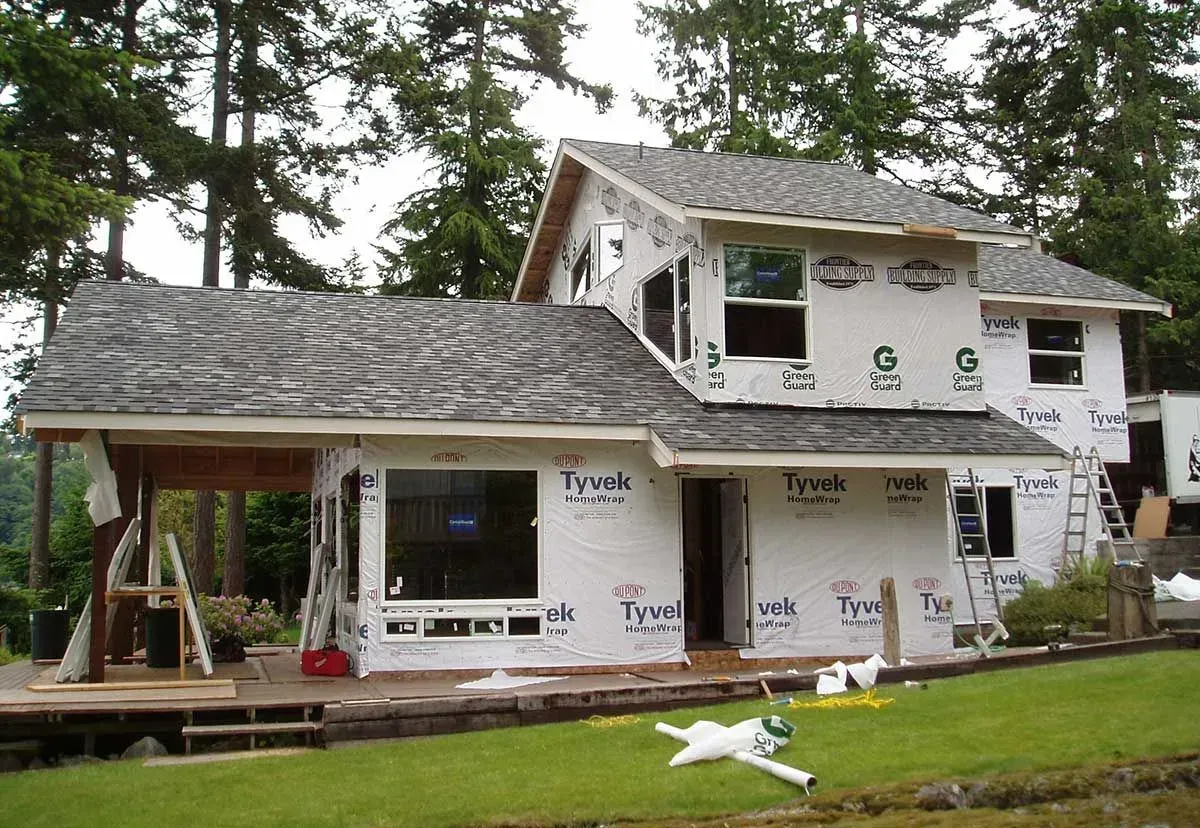 Two-story house under construction with white wrap, gray roof, and a front porch in a wooded yard