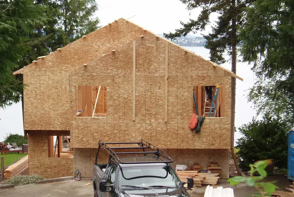 House under construction with OSB sheathing, a car parked in front, and trees in the background