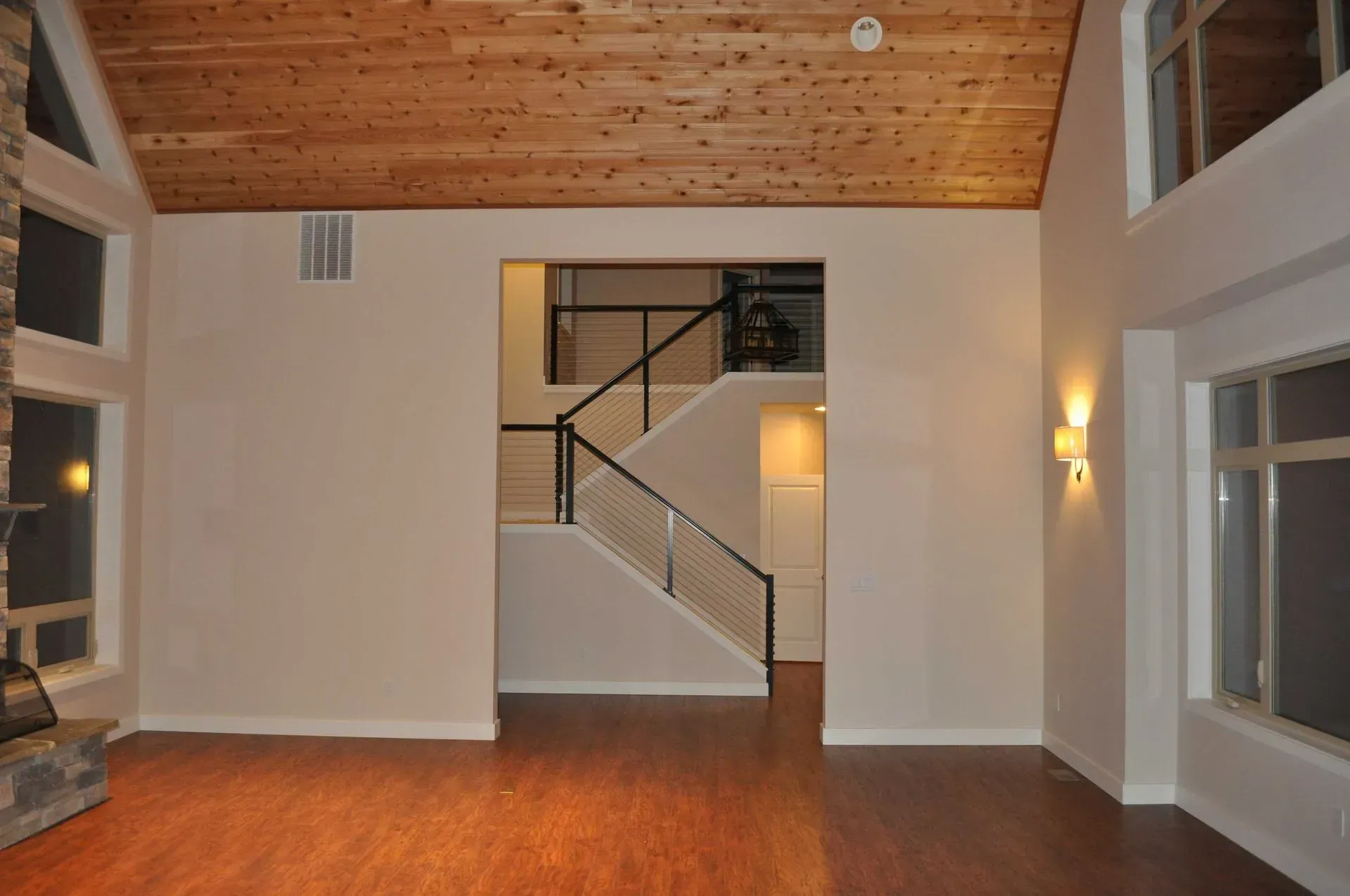 Spacious empty living room with hardwood floors, vaulted wood ceiling, and staircase in back.