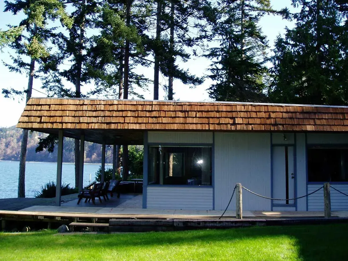 Small lakeside cabin with a wooden deck and chairs, surrounded by tall trees.