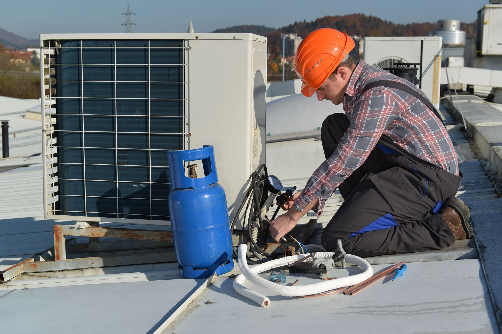 A worker in a hard hat kneeling on a rooftop, repairing an air conditioning unit next to a blue refrigerant tank.