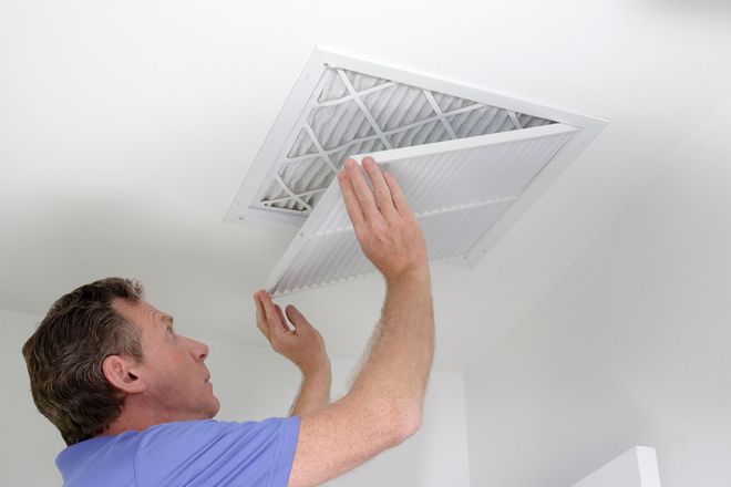 A person in a blue shirt replaces a pleated air filter in a white ceiling-mounted HVAC vent.