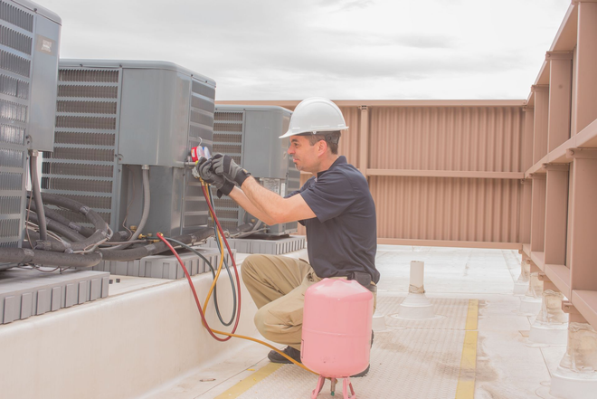 A worker wearing a hard hat and gloves inspects an HVAC unit on a rooftop, using gauges connected to a refrigerant tank.