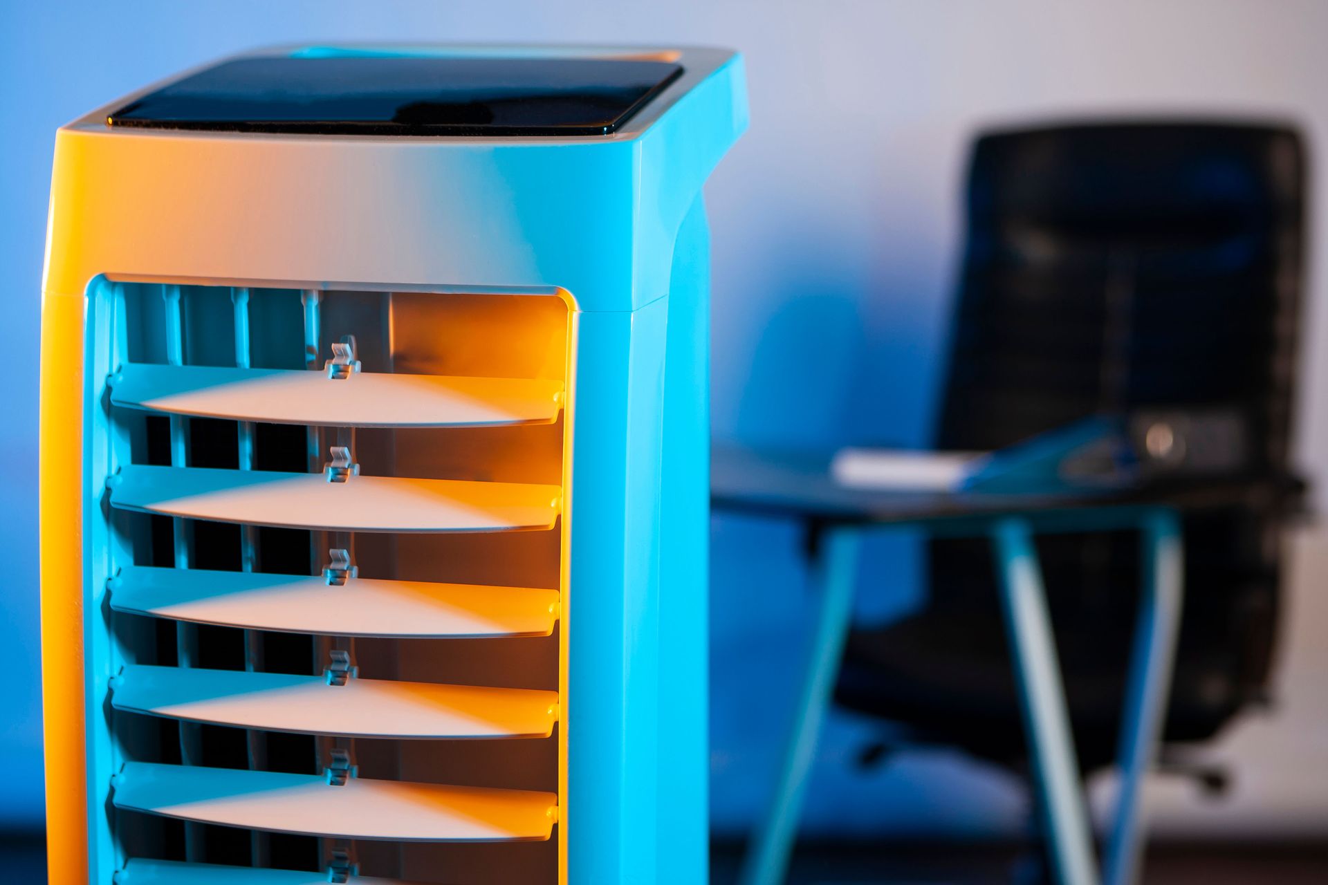 A portable air cooler with horizontal vents, illuminated by blue and orange light, sits in a blurred home office.