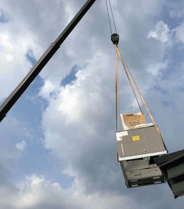 A crane lifts a large industrial HVAC unit through the air toward a building roof against a cloudy sky.