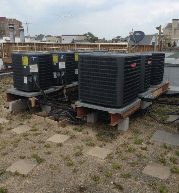 A row of dark grey HVAC condenser units on concrete pads and steel beams on a rooftop with sparse grass and pavers.