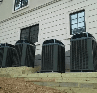 Four grey AC units sit on a tiered wooden platform against a light-colored building with two dark-framed windows.