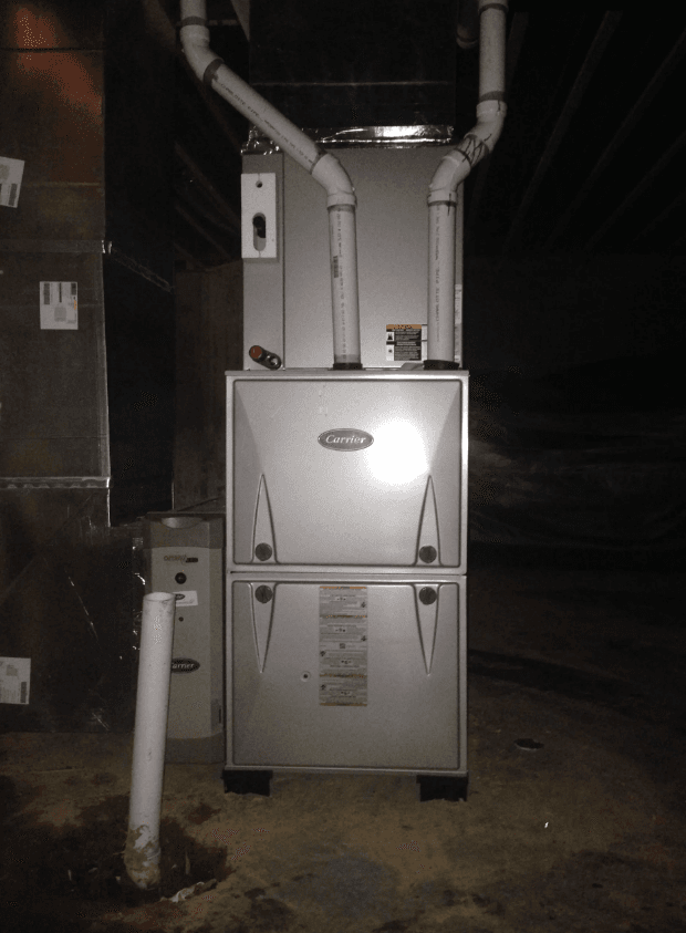 A gray furnace unit in a basement, featuring two white PVC pipes extending from the top and a vertical pipe on the floor.