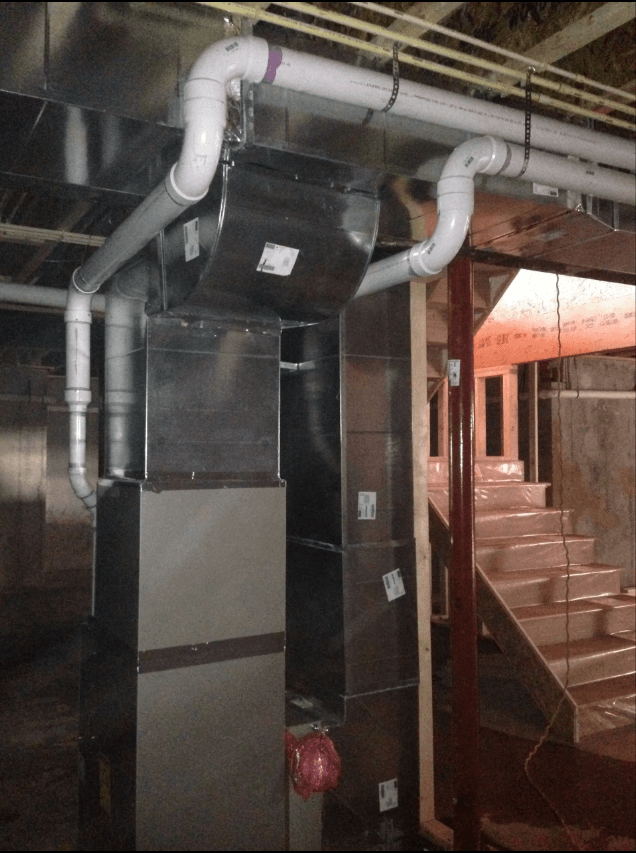 A furnace with white PVC exhaust piping and silver metal ductwork installed in an unfinished basement next to stairs.