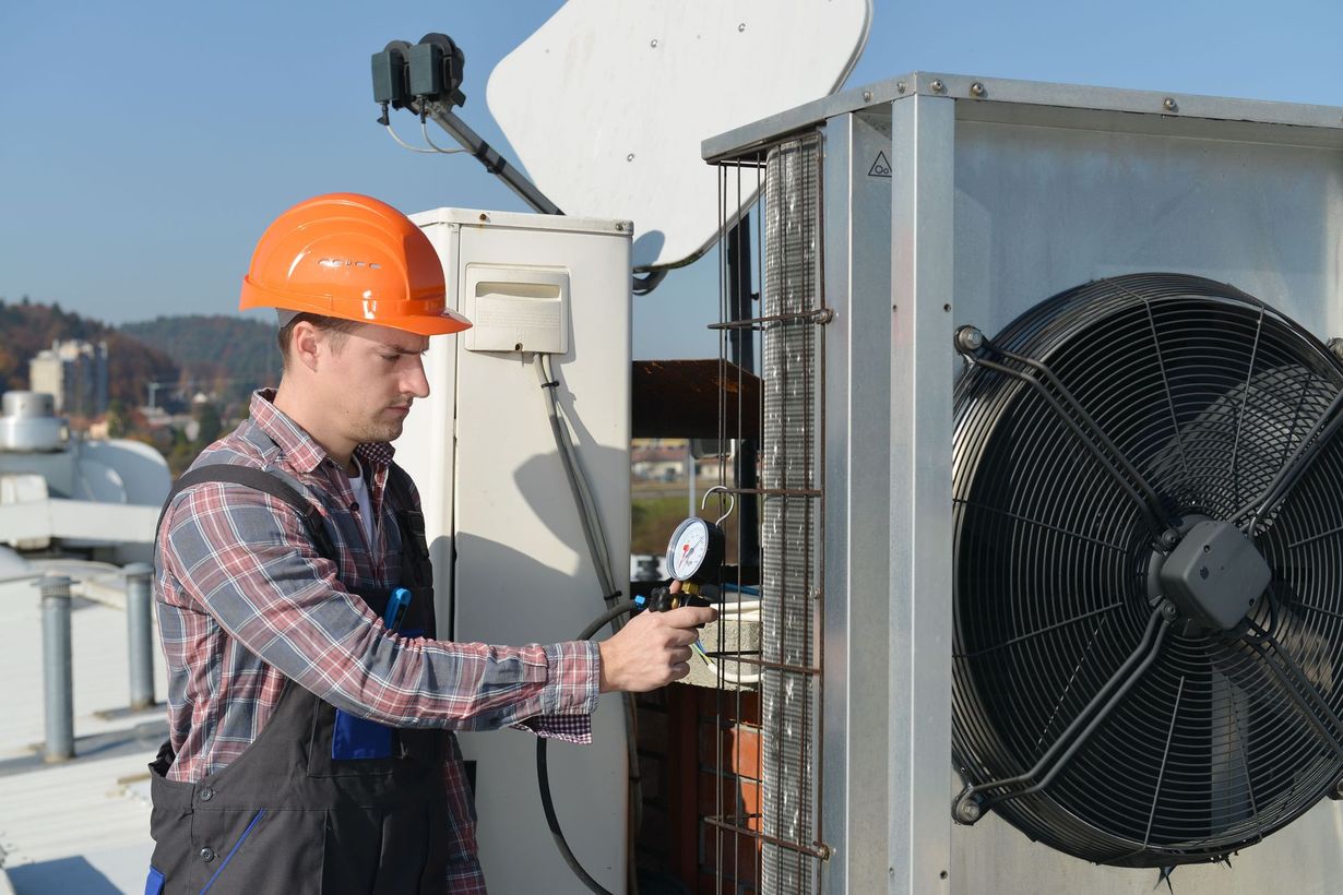 A worker in an orange hard hat and work overalls inspects an outdoor industrial air conditioning unit on a rooftop.