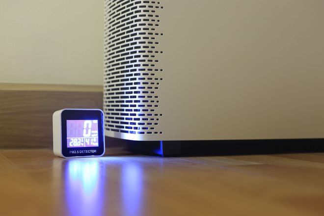A digital air quality monitor sits on a wood floor next to a white air purifier, displaying air quality metrics in blue.