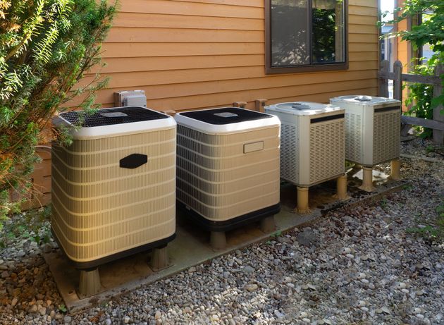 Four outdoor air conditioning condenser units sit on a gravel path against the side of a tan house.