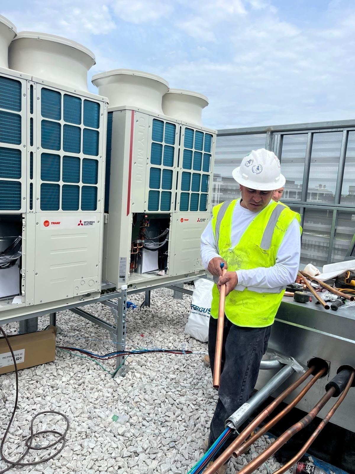 A worker in an orange hard hat and work overalls inspects an outdoor industrial air conditioning unit on a rooftop.