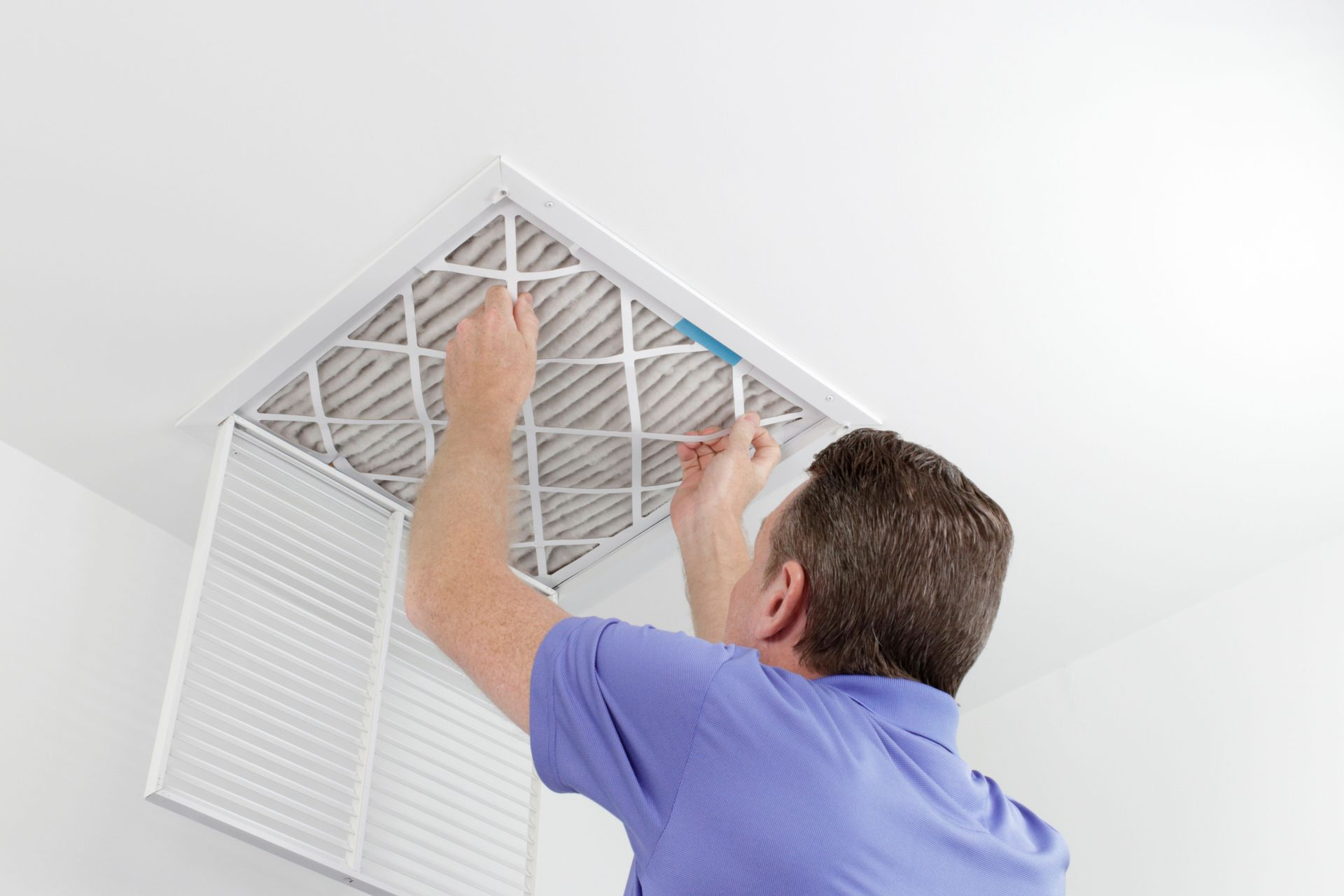 A person in a blue shirt replaces a pleated air filter in a white ceiling vent.