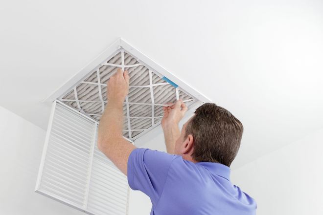 A person in a blue shirt replaces a pleated air filter in a white ceiling vent.