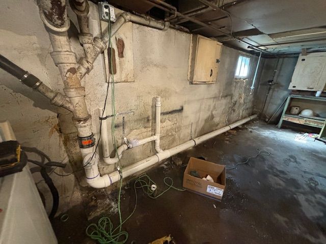 Basement with exposed pipes, wet floor, and concrete walls. A cardboard box sits on the floor near a small window.