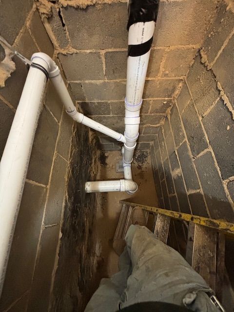 White PVC pipes in a dark basement with cinder block walls. One pipe runs diagonally, a person's legs and stairs are visible.