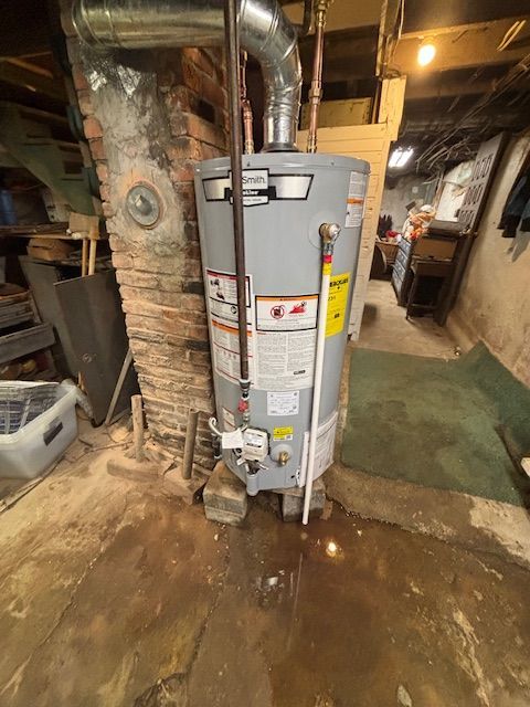 Water heater in a basement, with wet floor. Grey tank, brick wall, silver vent.