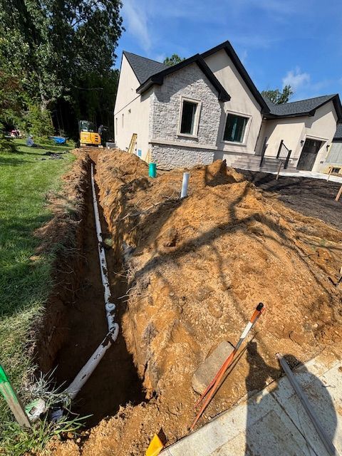 A trench in a yard with exposed white pipes, next to a beige house, under a blue sky.
