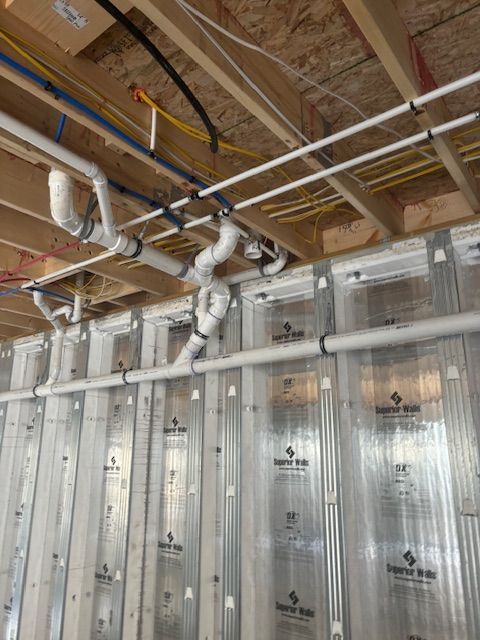 Pipes and wiring run along a wooden ceiling. Pipes are white, wires are yellow and blue, and the wall is framed.