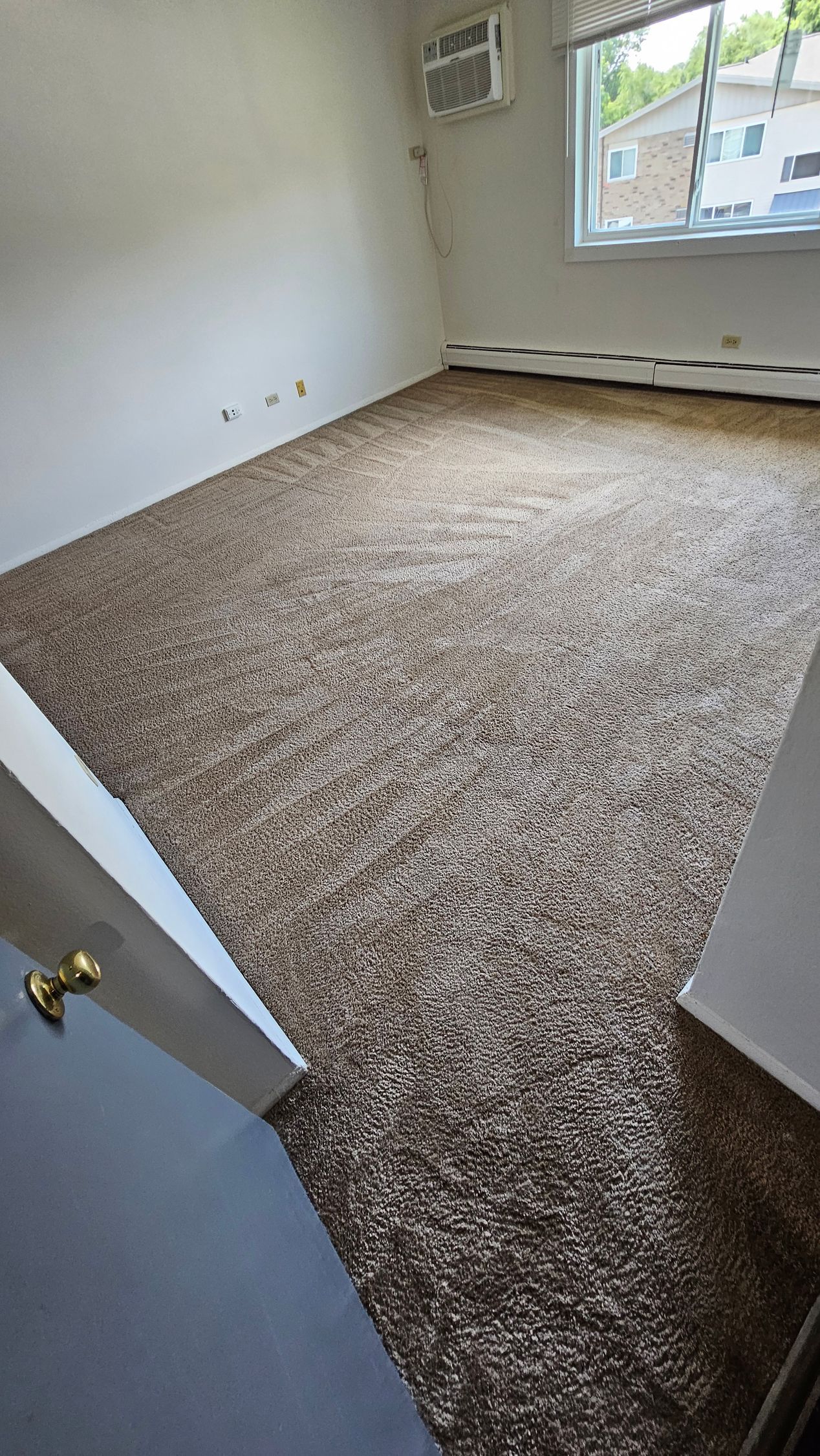 Empty beige-carpeted bedroom with white walls and a window, viewed from the doorway.
