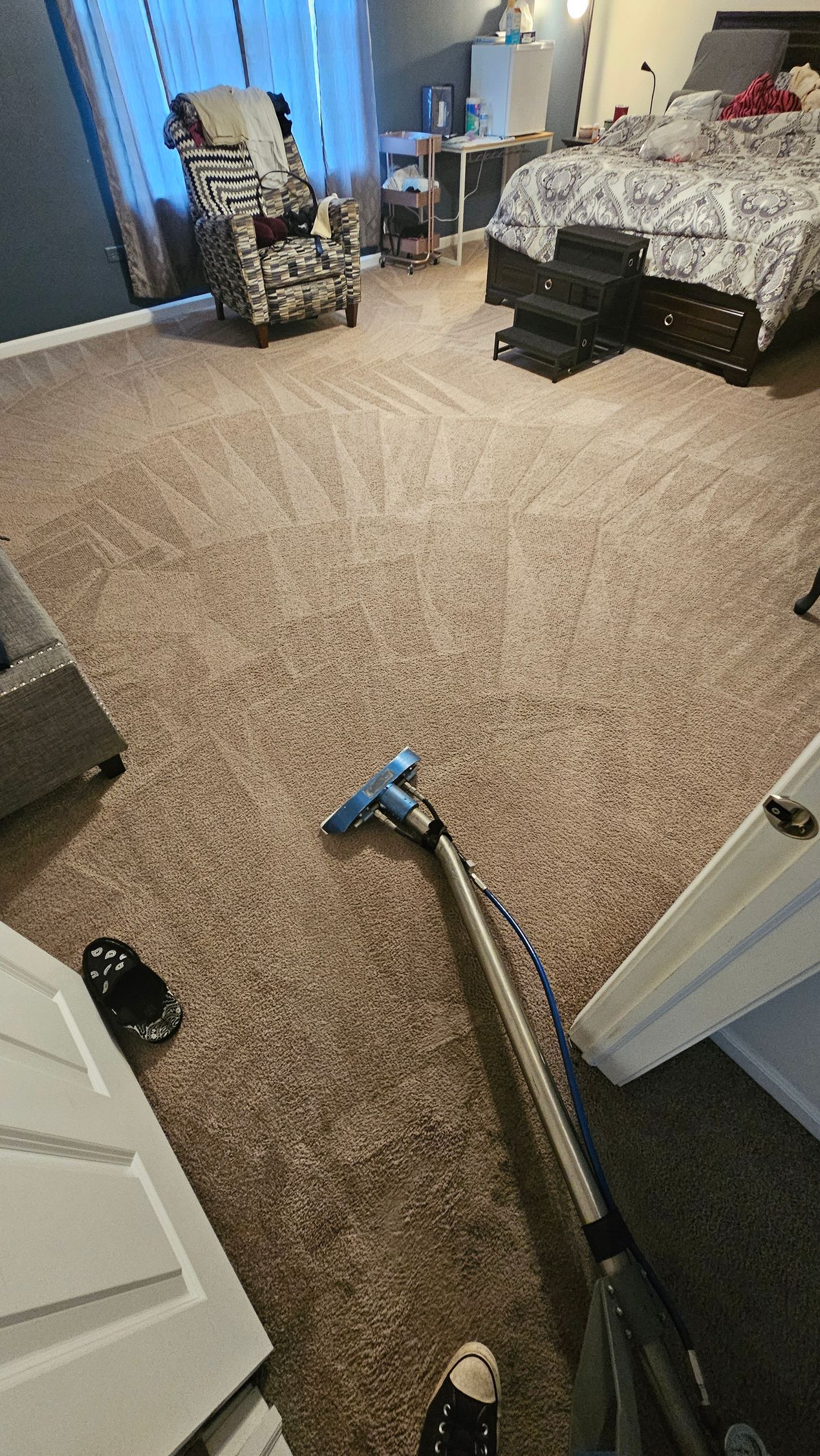 Vacuum cleaner on beige carpet in a cluttered bedroom with a bed and chair.