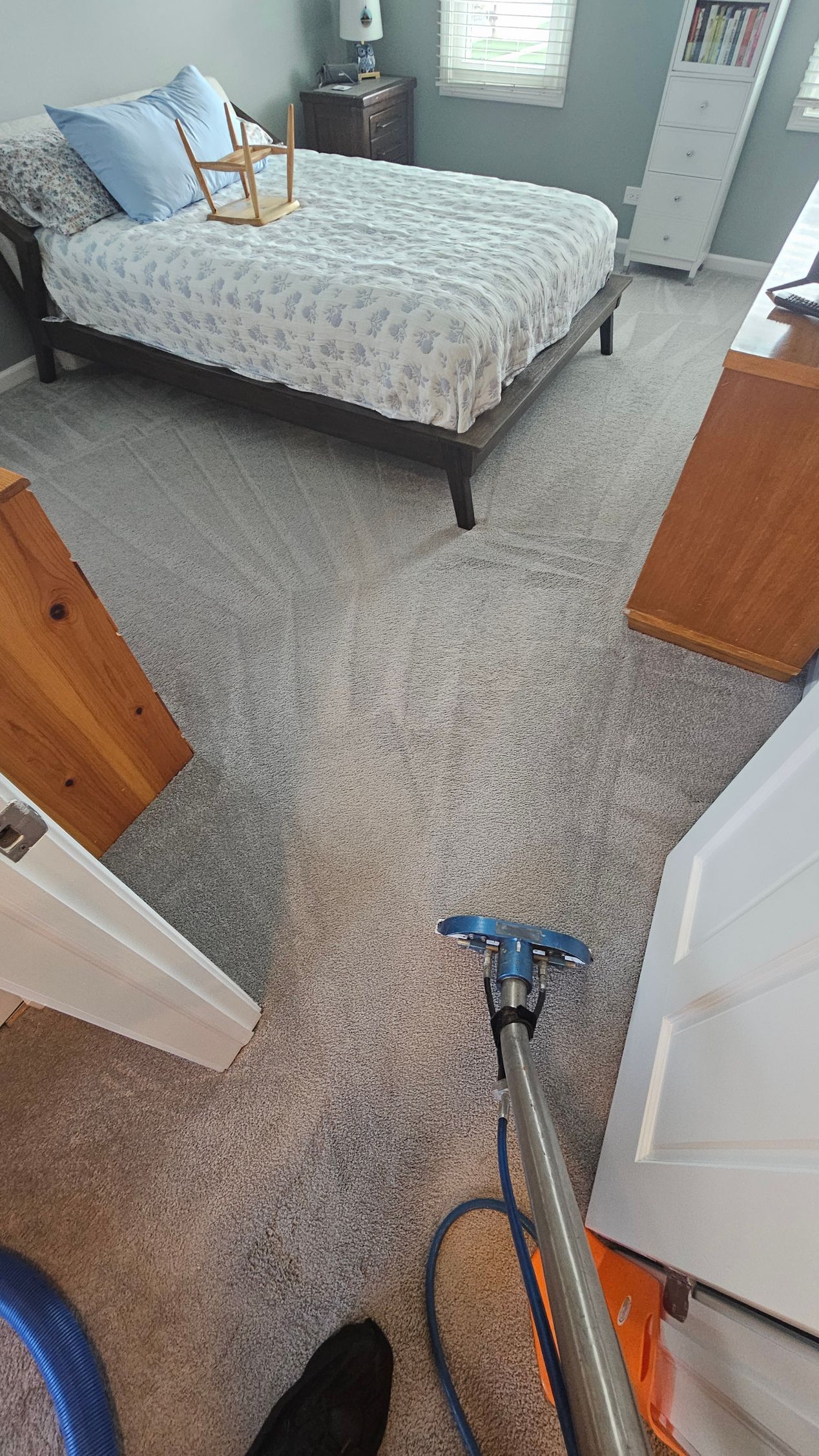 Bedroom with bed, dresser, and vacuum on speckled carpet, viewed from a doorway.