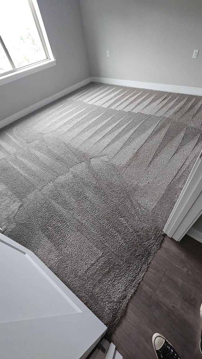 Empty gray-carpeted room with a window, white walls, and doorway view into a hall.