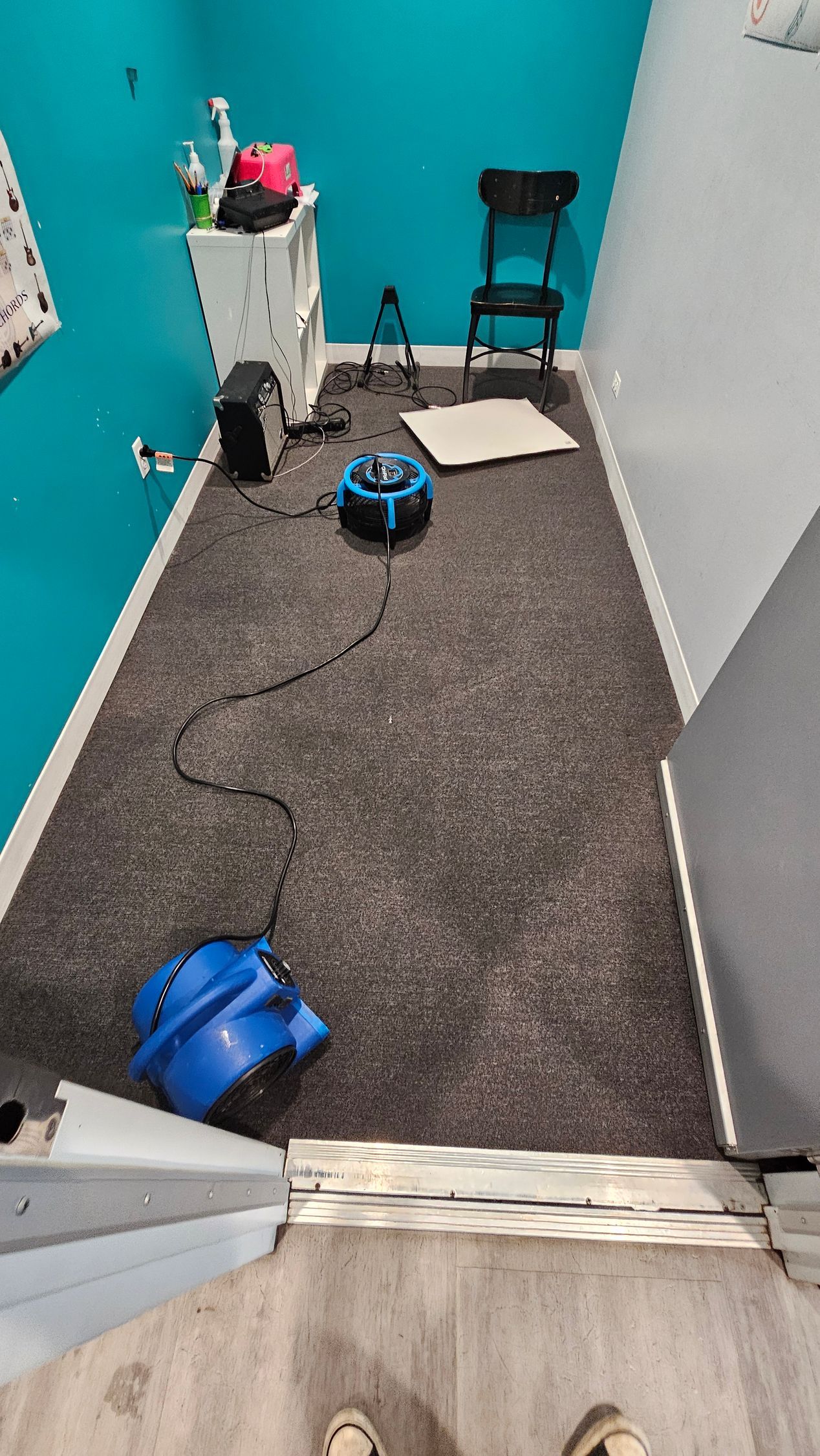 Small room with teal walls, carpet, a chair, and several blue cleaning machines with cords on the floor.