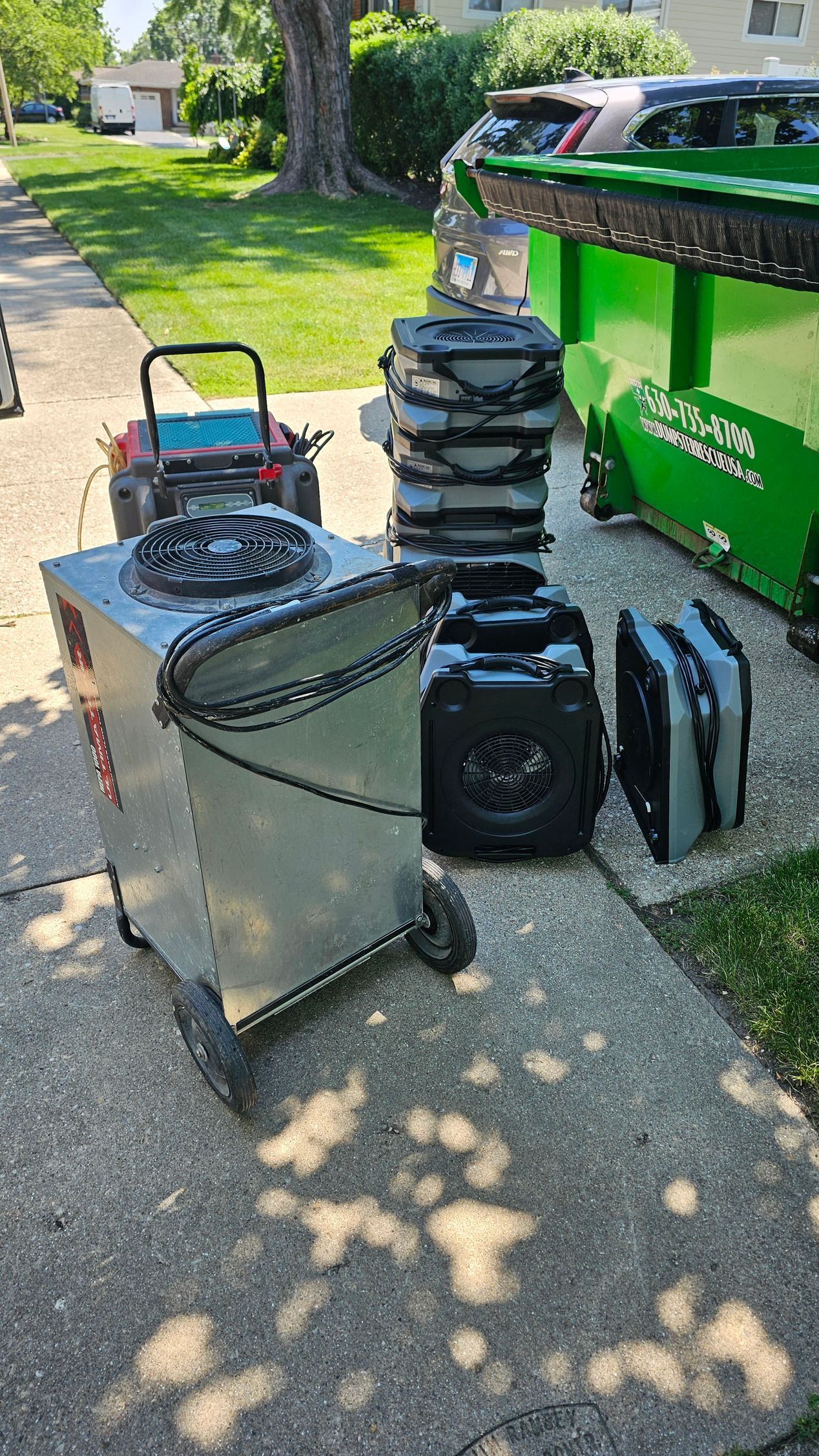 Wheelbarrow with coiled hose, stacked black containers, and a green recycling bin on a sunny sidewalk.