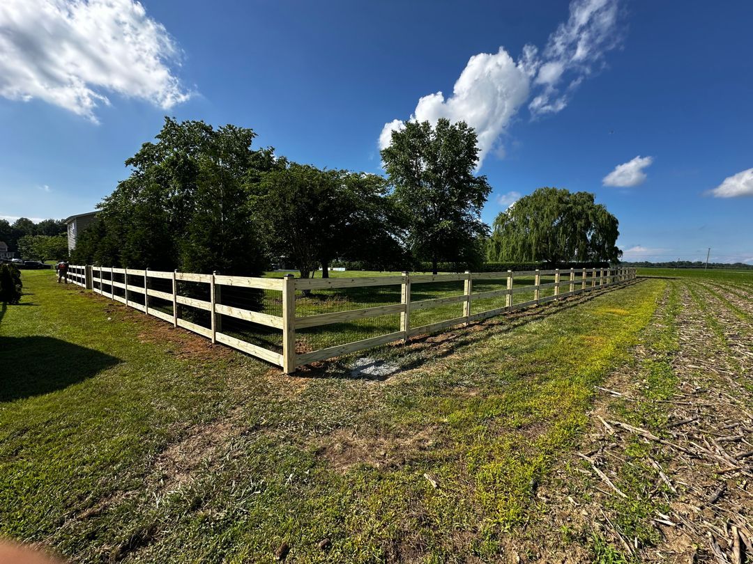 A wooden fence encloses a grassy area with trees, set against a blue sky with clouds.
