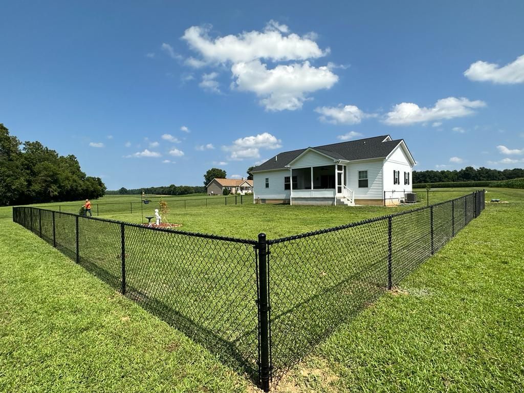 Black chain-link fence surrounds a white house with a porch. Two people work in the yard on a sunny day.