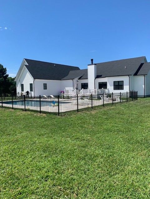 White house with black roof, swimming pool, fenced yard, and green grass on a sunny day.