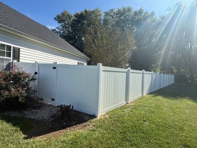 White vinyl fence borders a house, with green grass and trees in the sunny outdoor setting.