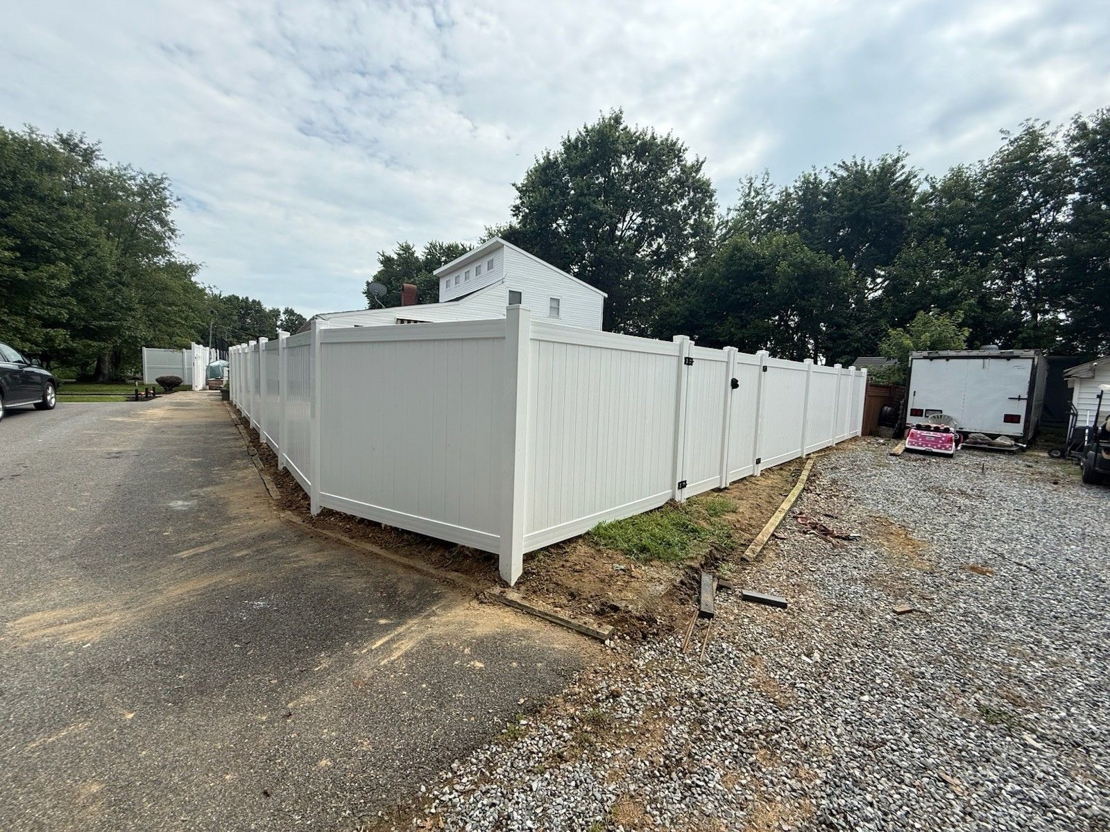 White vinyl fence surrounds a gravel lot with a building, trees, and vehicles in the background under a cloudy sky.