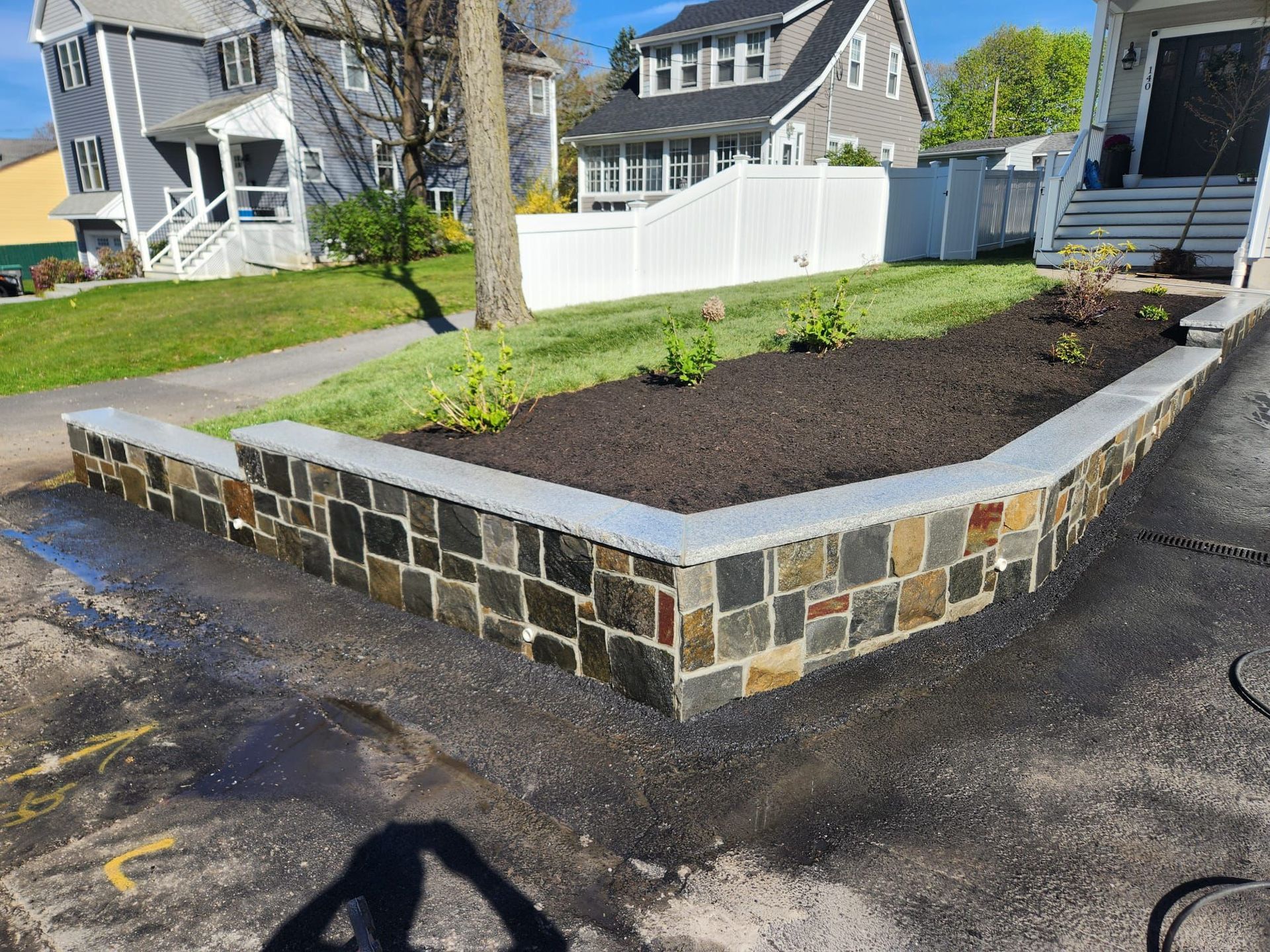 Raised stone planter bed with fresh mulch and a small tree beside a driveway in a suburban yard