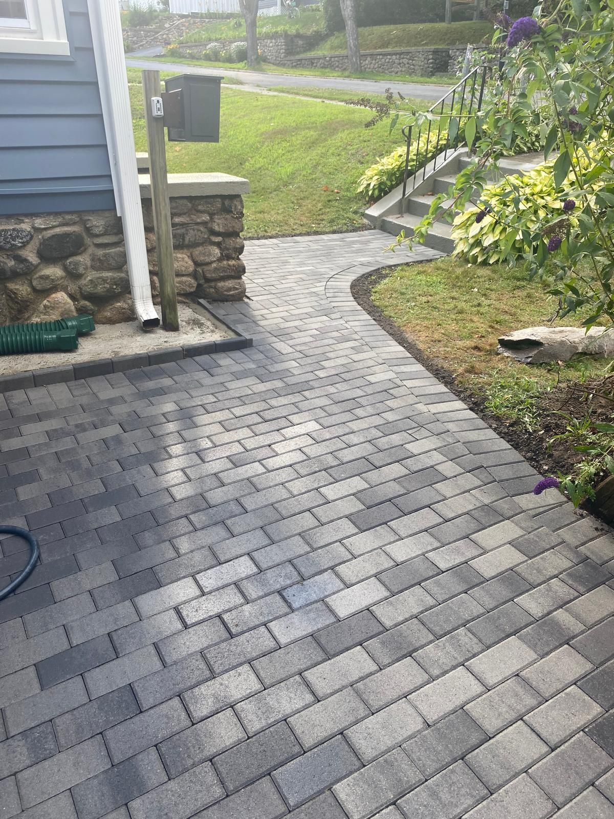Curved gray paver walkway beside a house, leading through a landscaped yard with grass and shrubs.