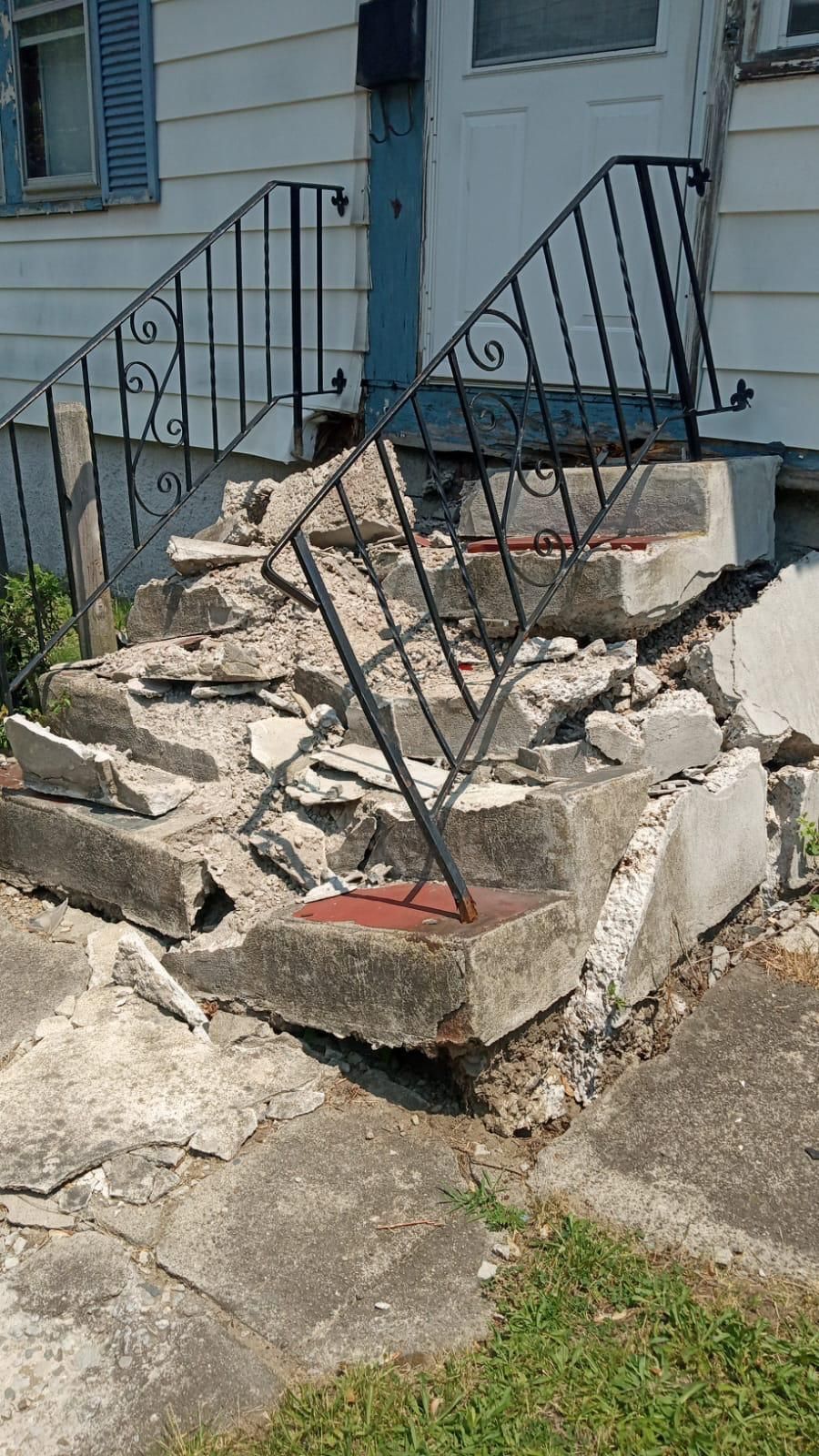 Broken concrete porch steps with a bent black metal railing outside a house