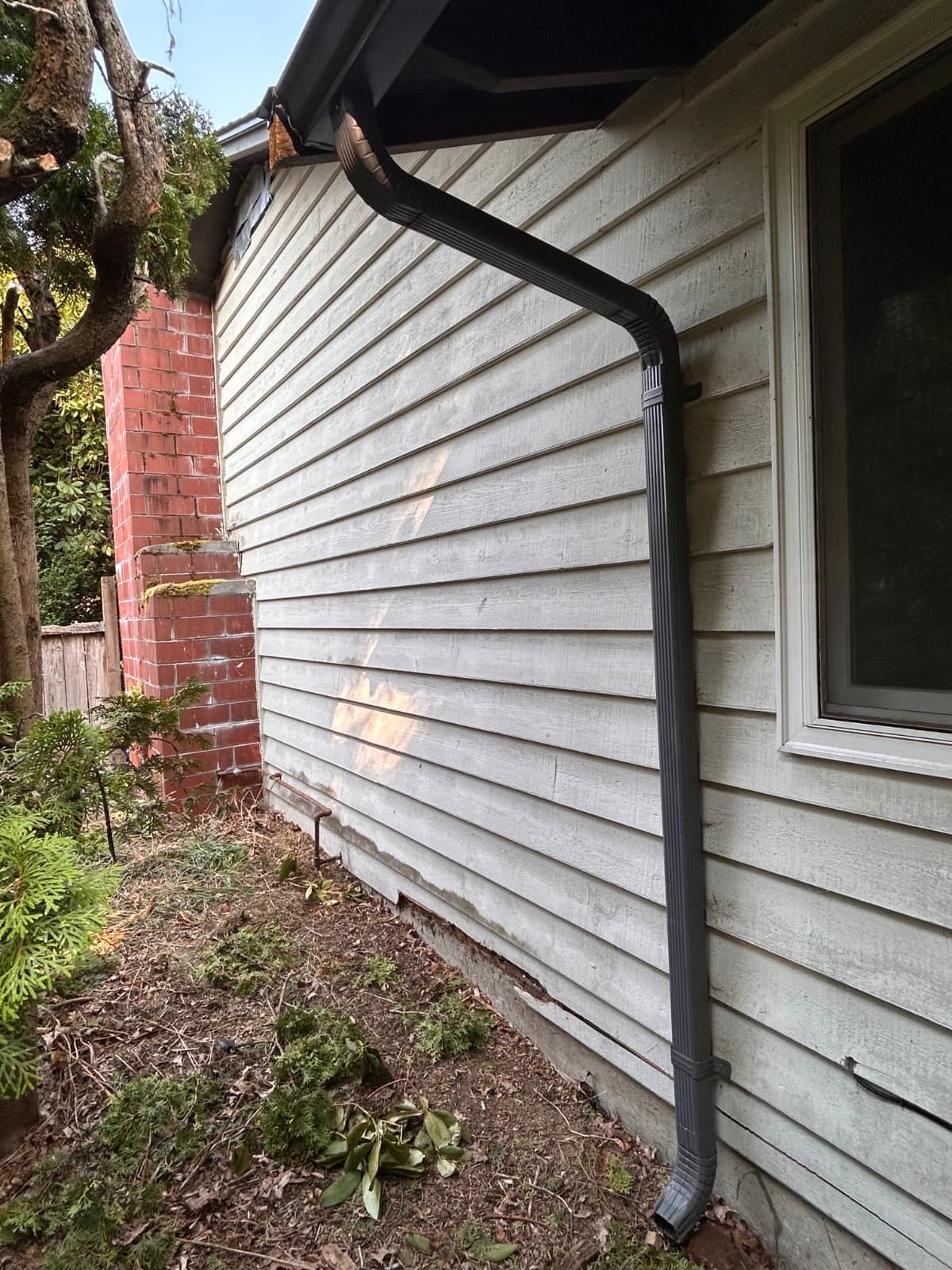 Damaged white house siding with a bent downspout beside a window and red brick wall