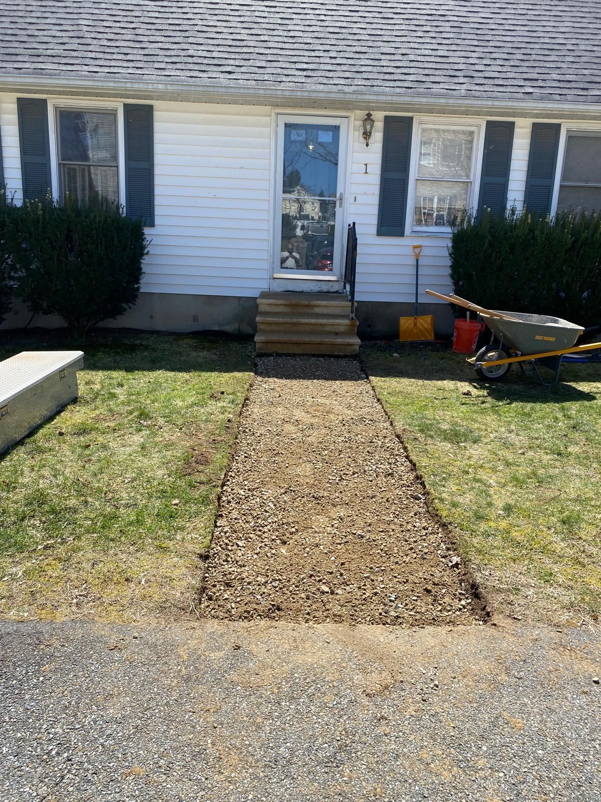 Front walkway leading to a white house entry with steps, shrubs, and a wheelbarrow on the right.