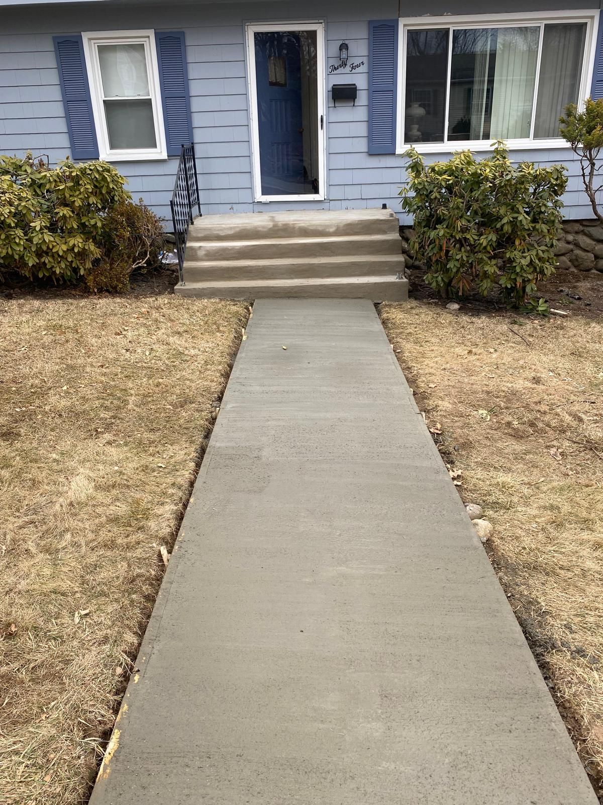 Concrete walkway leading to a blue house with steps and a front door, flanked by shrubs and dry grass
