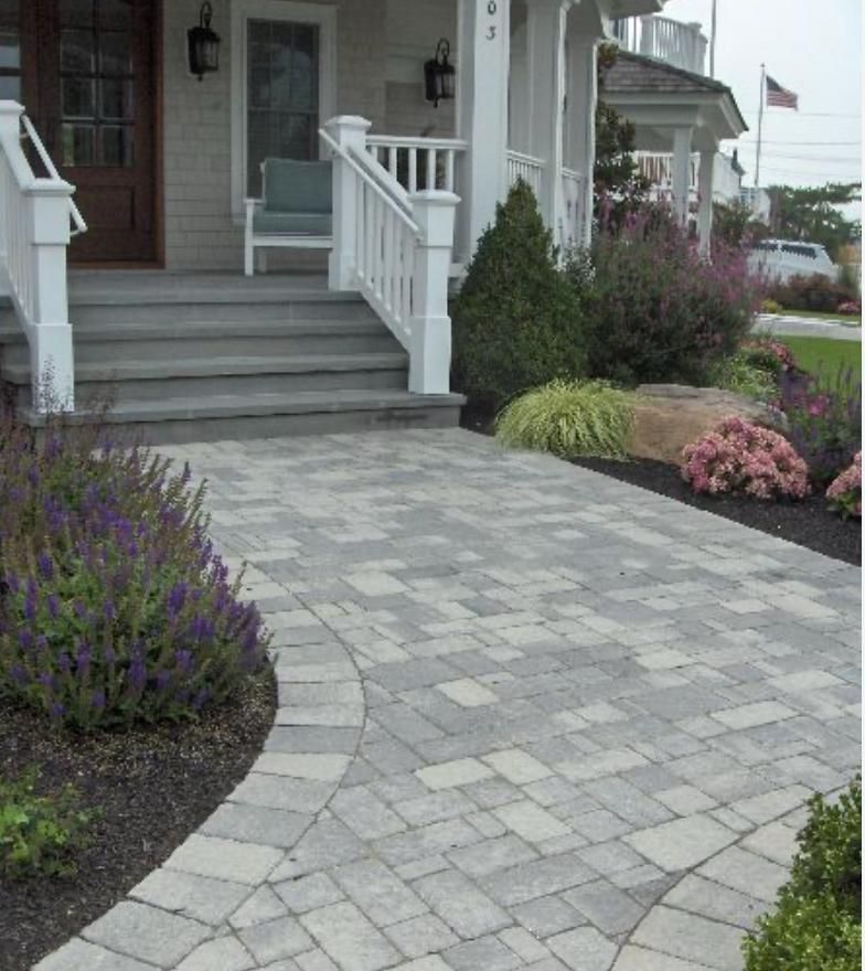 Stone-paved front walkway leading to a house entrance with white steps, railings, and flower beds